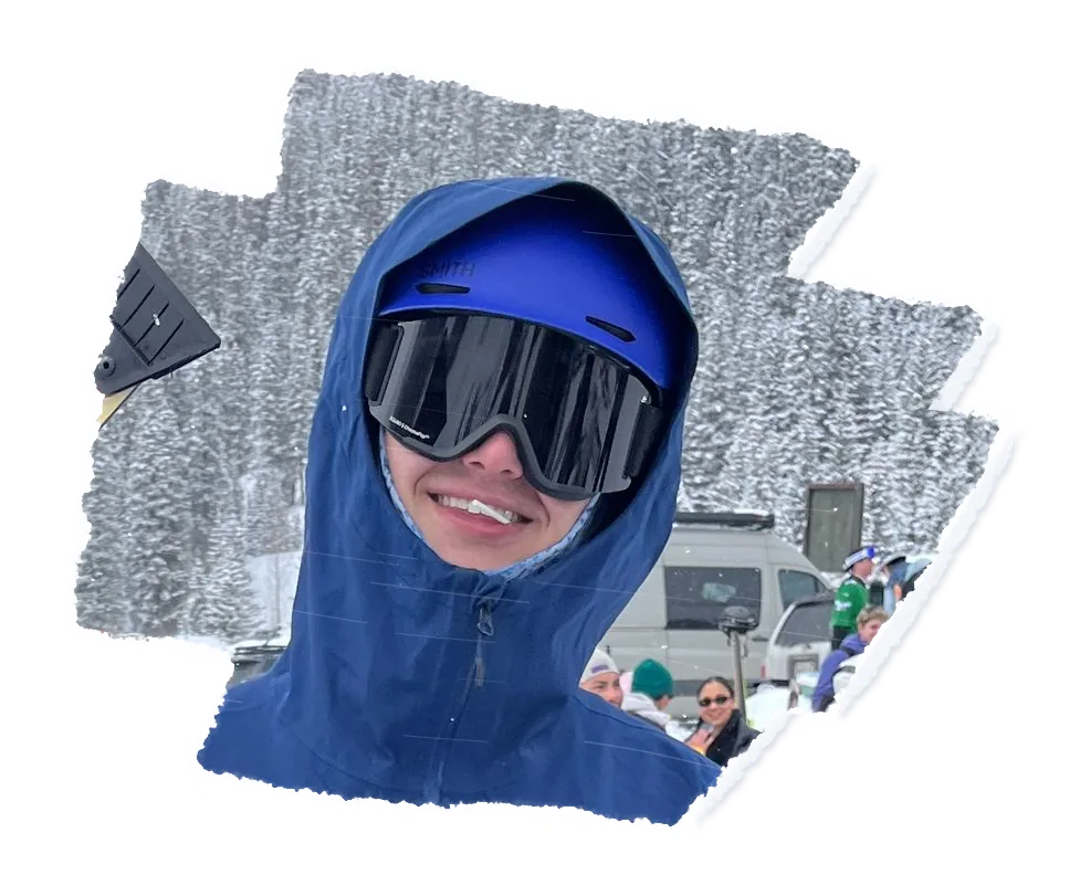 A skier in a blue hood and dark goggles smiling, snowy pines behind them.