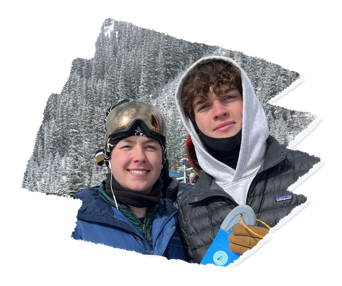 Two people at a resort base in falling snow, one in goggles and one in a hood, a blue scraper in a gloved hand.