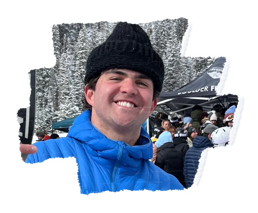 A person in a black beanie and a blue jacket grinning, a crowd at a resort base behind them.