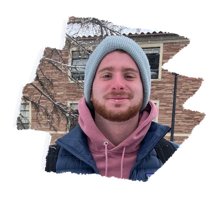 Someone in a grey beanie and a pink hoodie smiling, brick halls behind them.