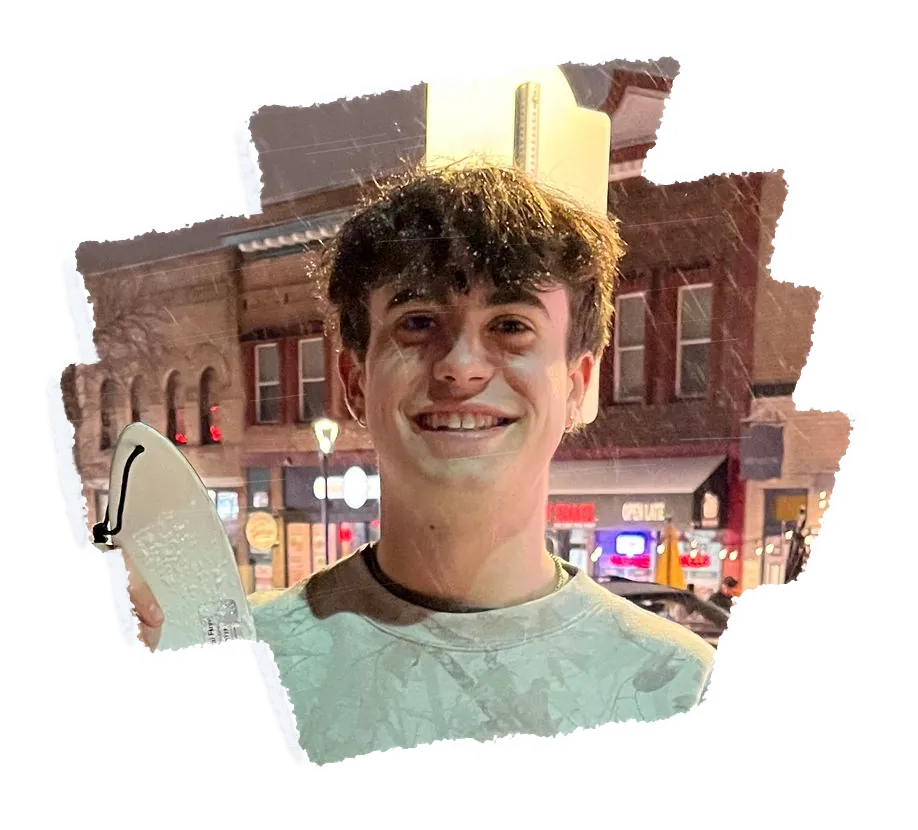 A person smiling in falling snow at night, a scraper at their shoulder and a lit street behind.