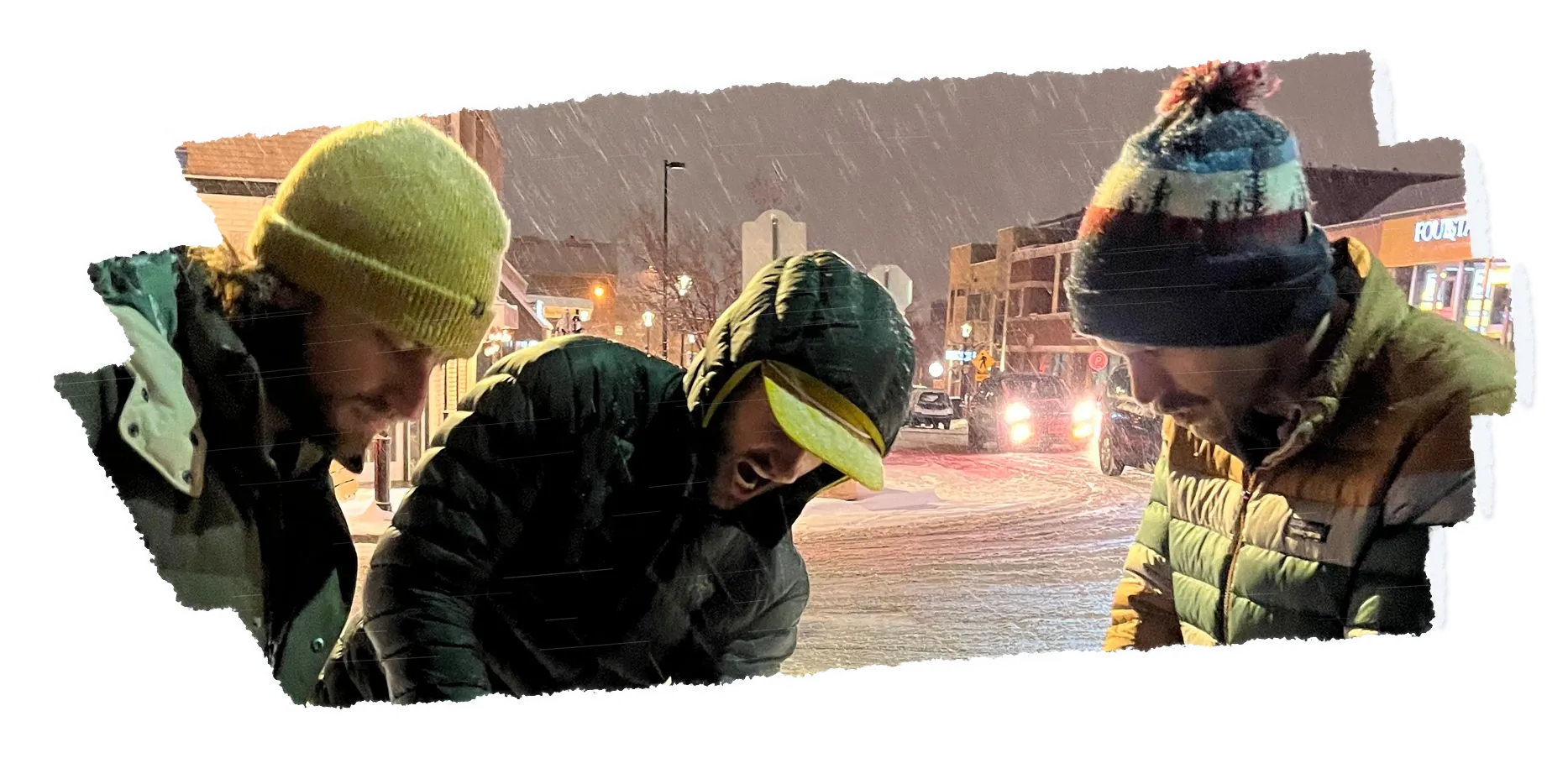 Three people in beanies leaning over a table in heavy snow at night, looking down.