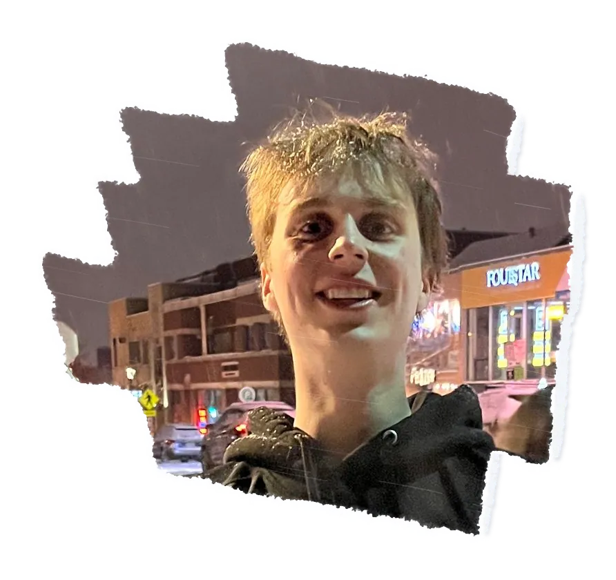 A person smiling up at the camera at night, shop fronts lit behind them.