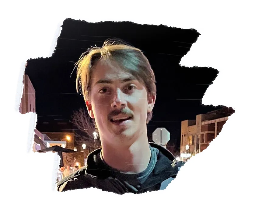 A person with a moustache looking into the camera, a lit street at night behind them.