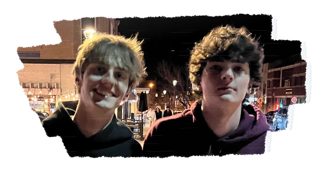 Two people standing shoulder to shoulder at night, lit shop fronts behind them.