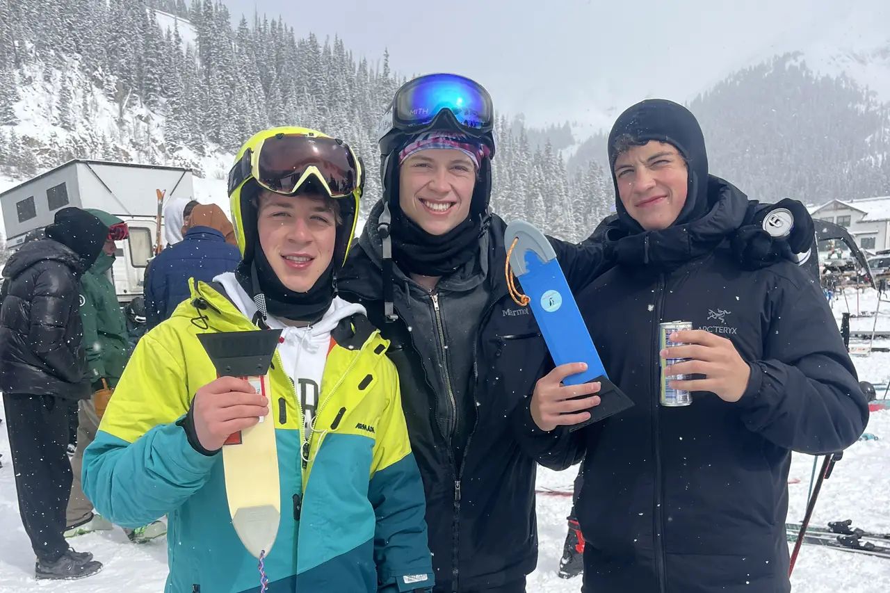 Three skiers in ski gear standing in the snow with drinks and a scraper.