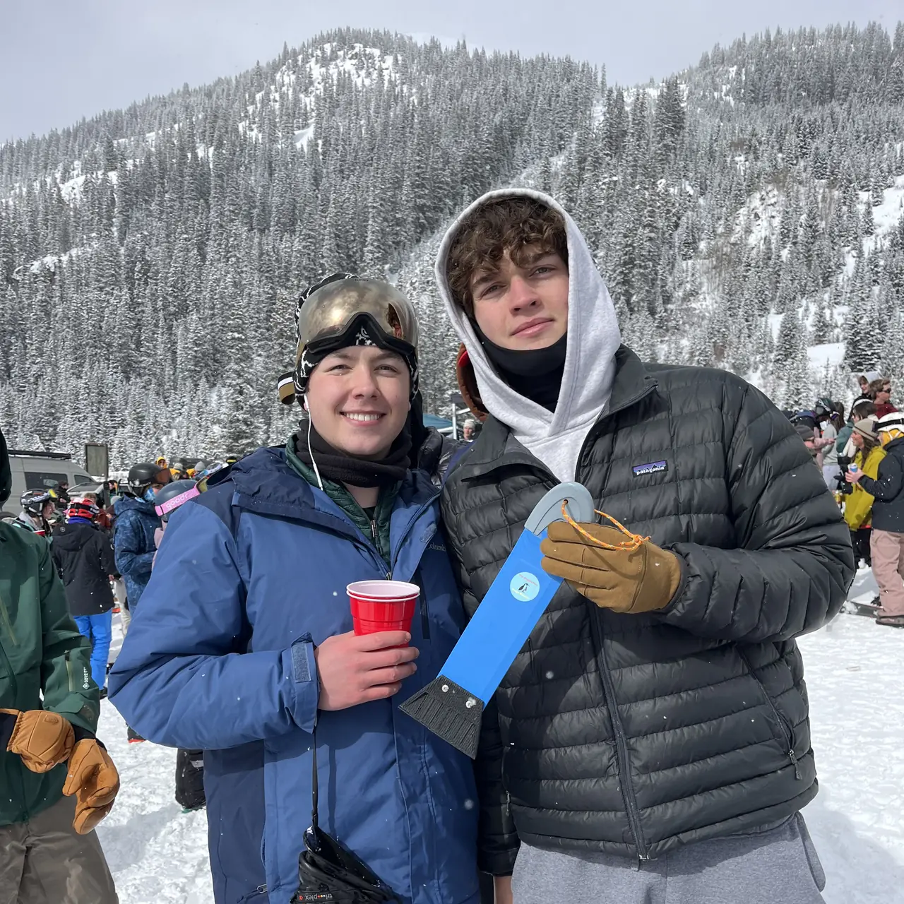 Two people at a resort base in falling snow, one with a scraper and one with a cup.