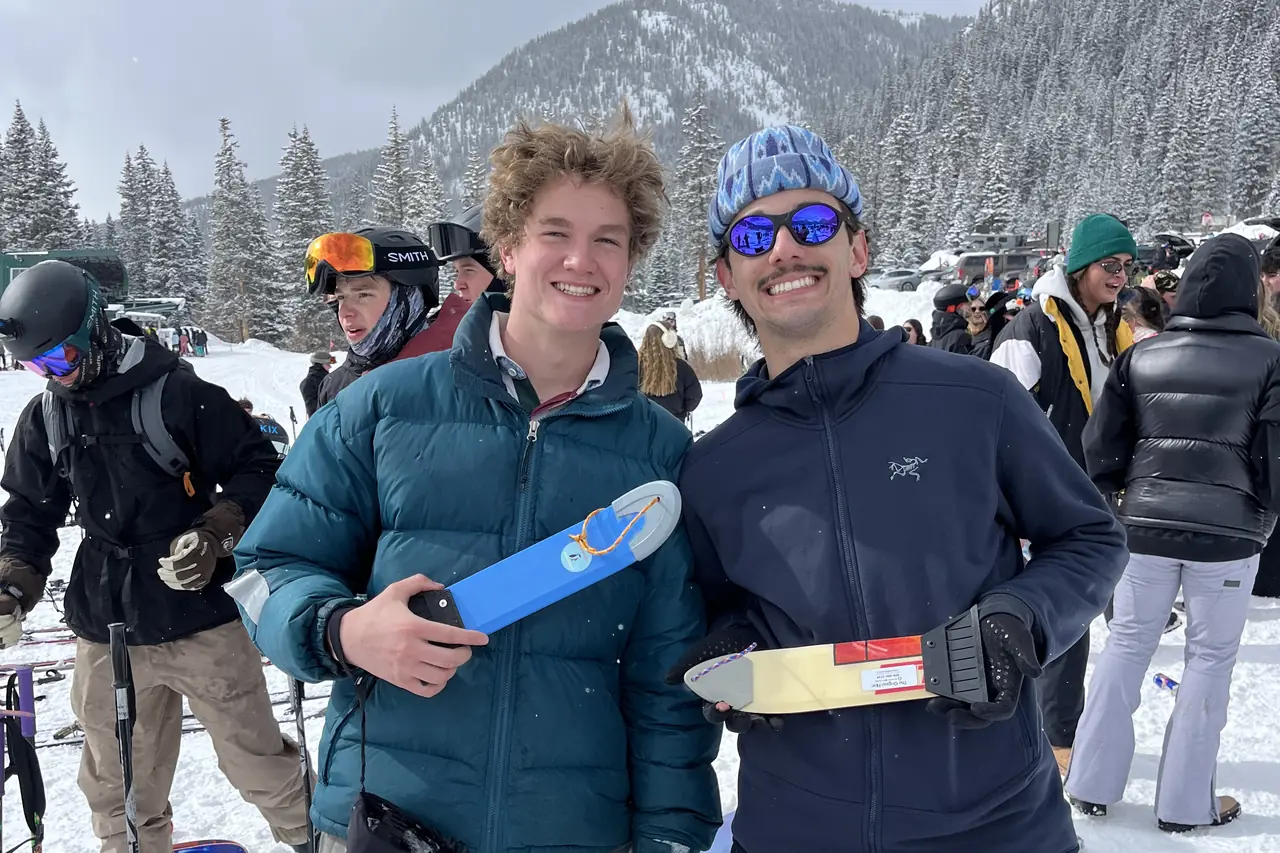 Two snowboarders at a resort base holding scrapers, skis stacked behind them.