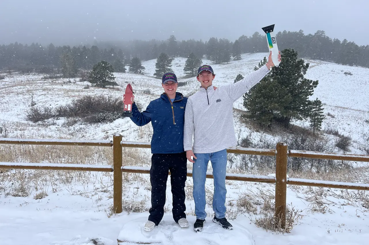 Two people on a snow-covered hillside in fog, arms raised, scrapers in hand.
