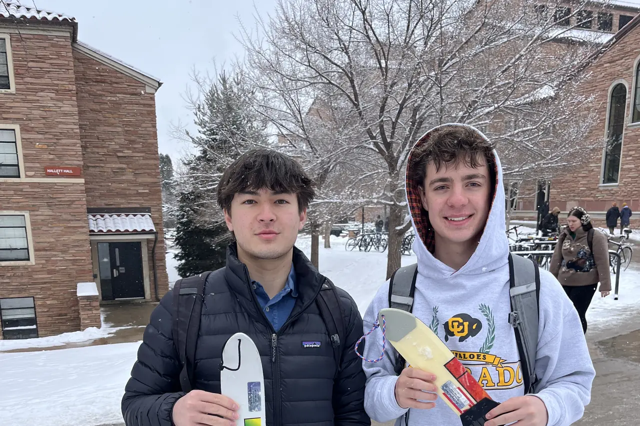 Two people standing behind a table on a snowy campus path, holding scrapers.