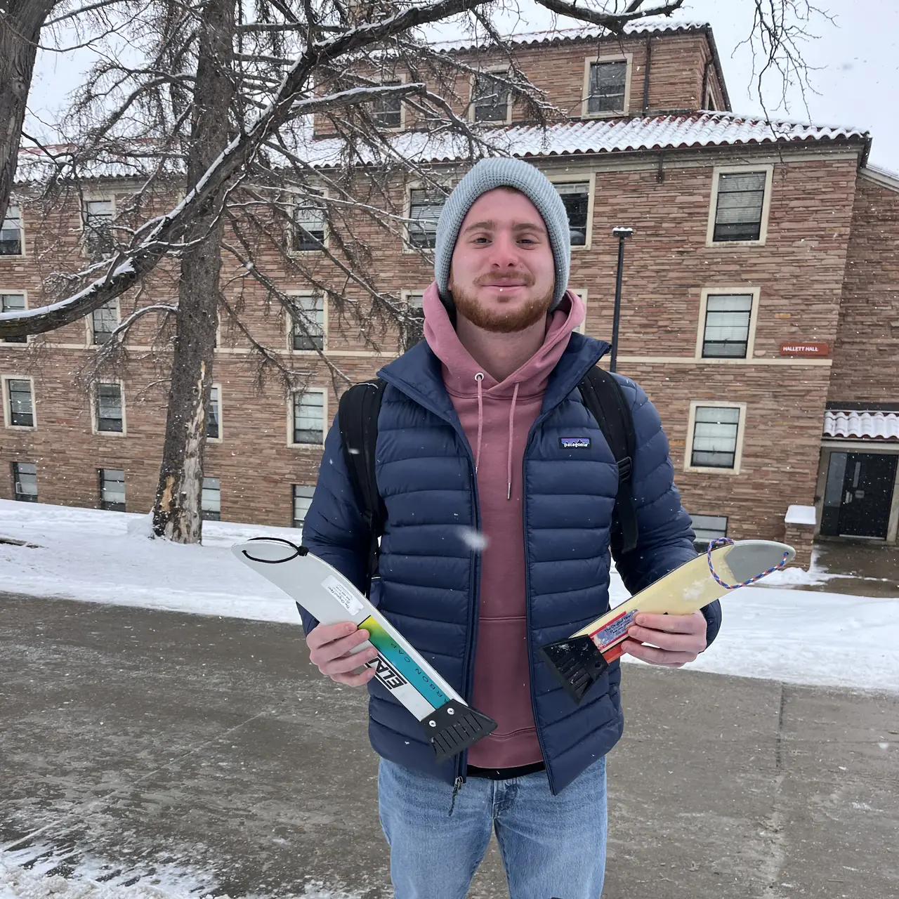 Someone in a beanie holding two scrapers over a table in the snow, brick halls behind.
