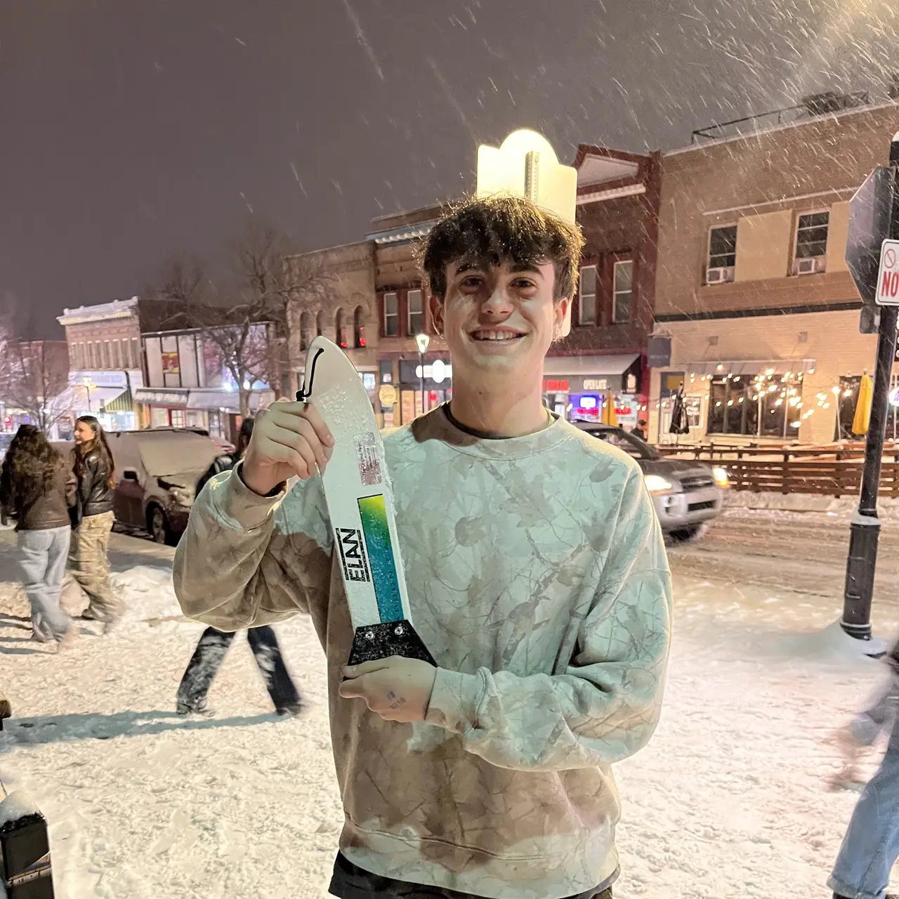 A person holding a scraper up in falling snow, a lit street behind them.