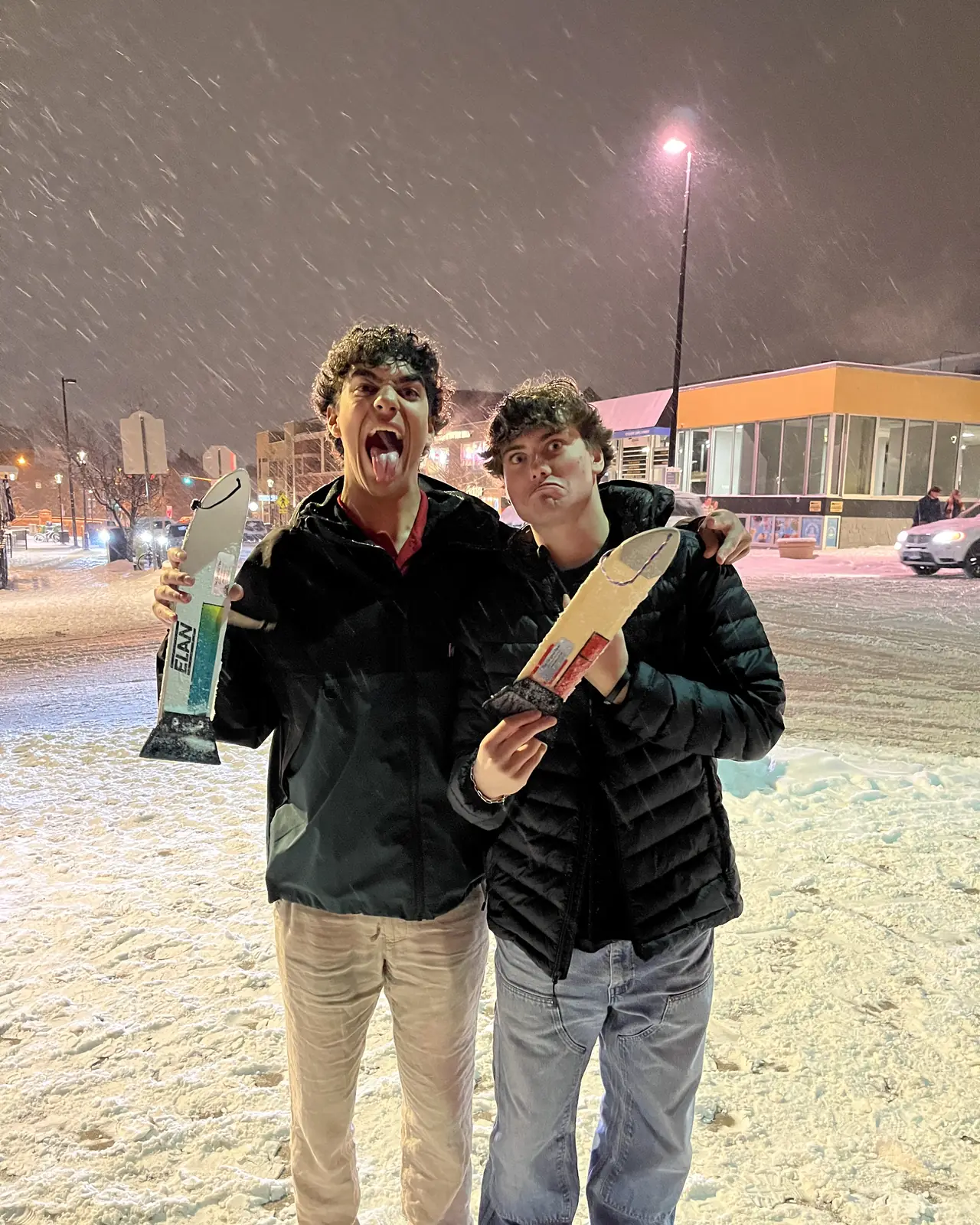 Two people pulling faces in falling snow under a street light, a scraper in each hand.