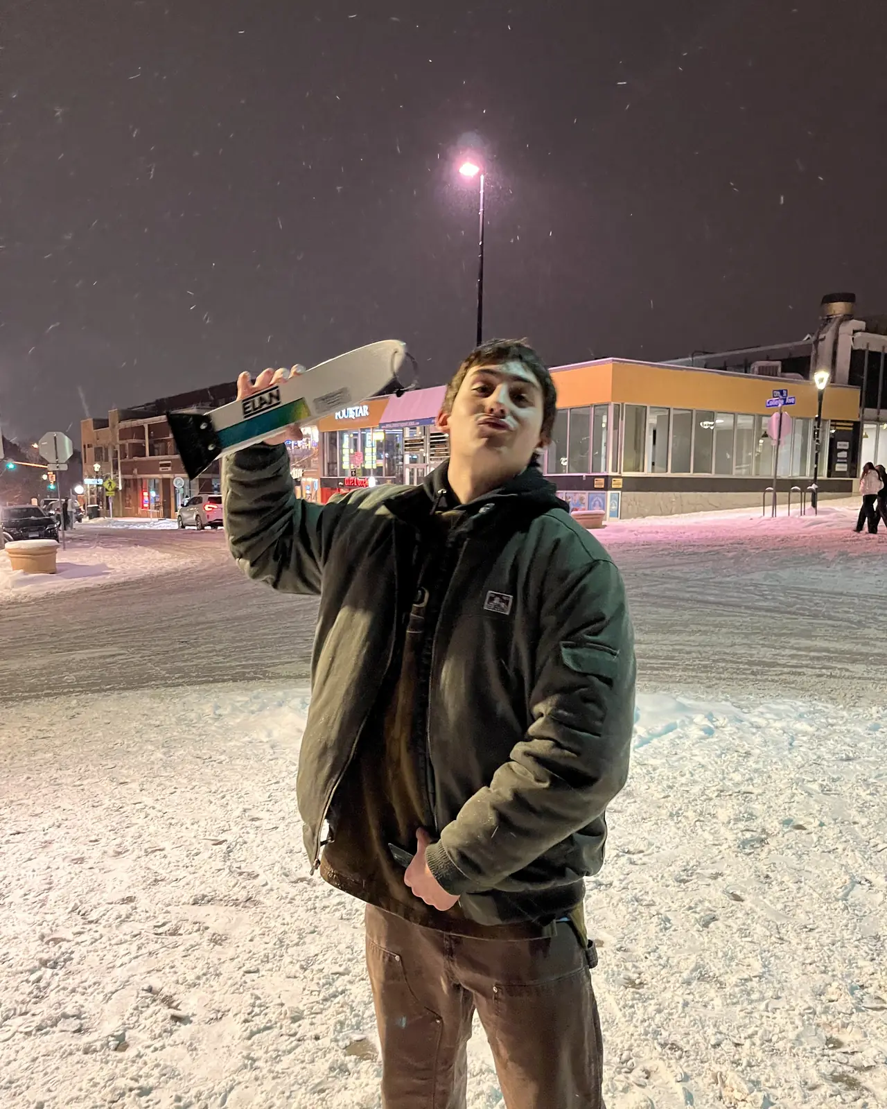 A person holding a scraper out at arm's length on a snowy street at night.