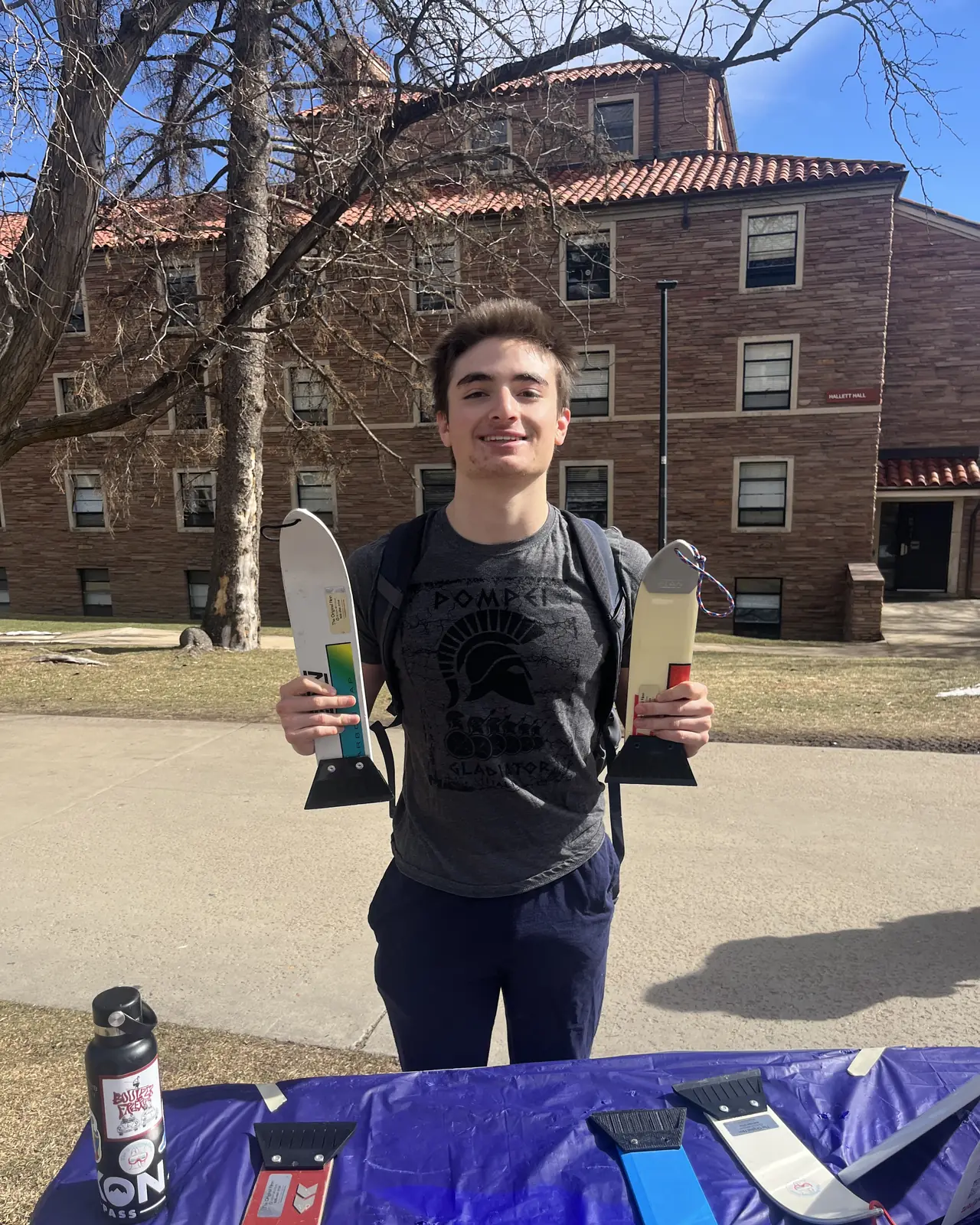 Someone with a backpack holding a scraper in each hand on a campus path.