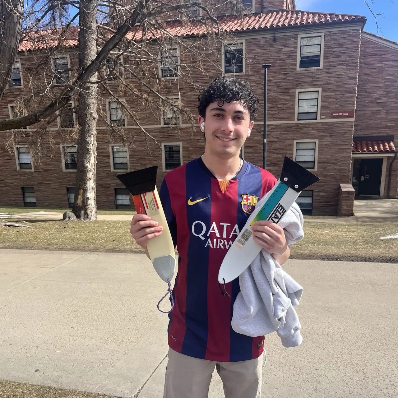 Someone in a football shirt holding two scrapers, low winter sun across a brick building.