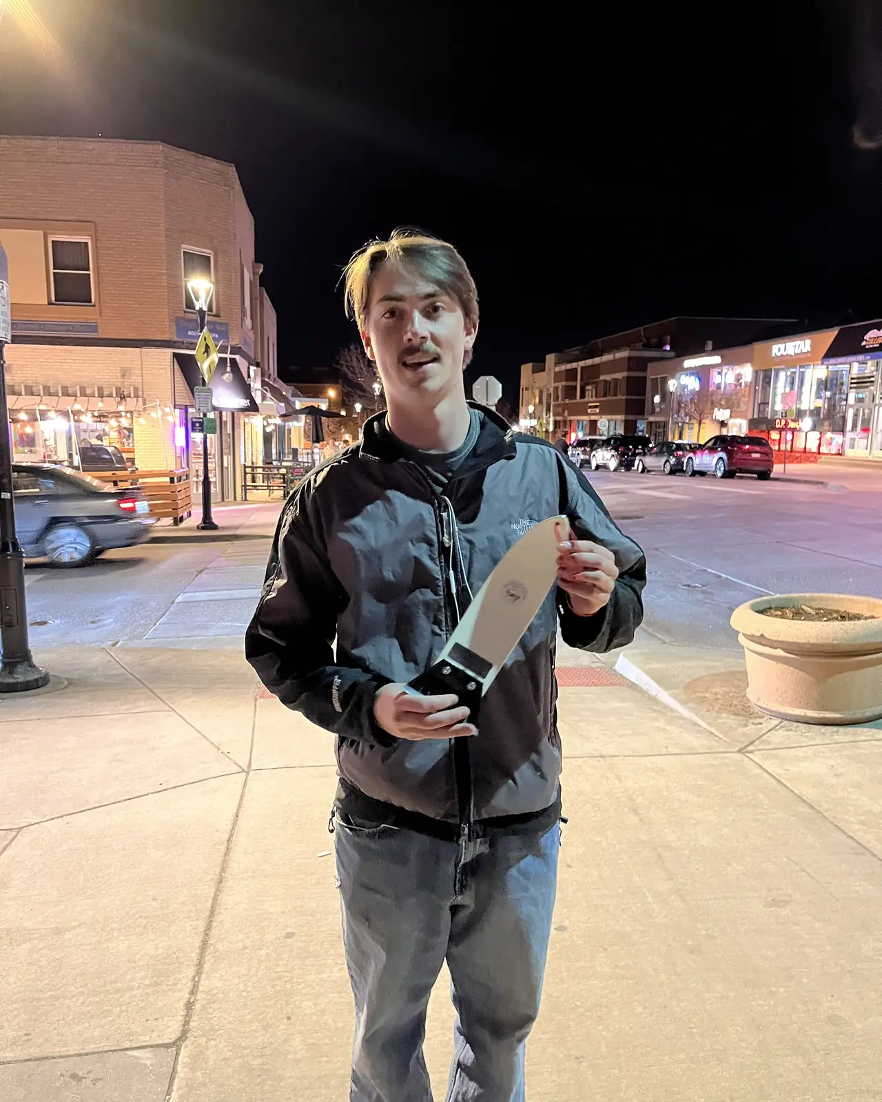 A person standing alone on a lit pavement at night, holding a scraper.